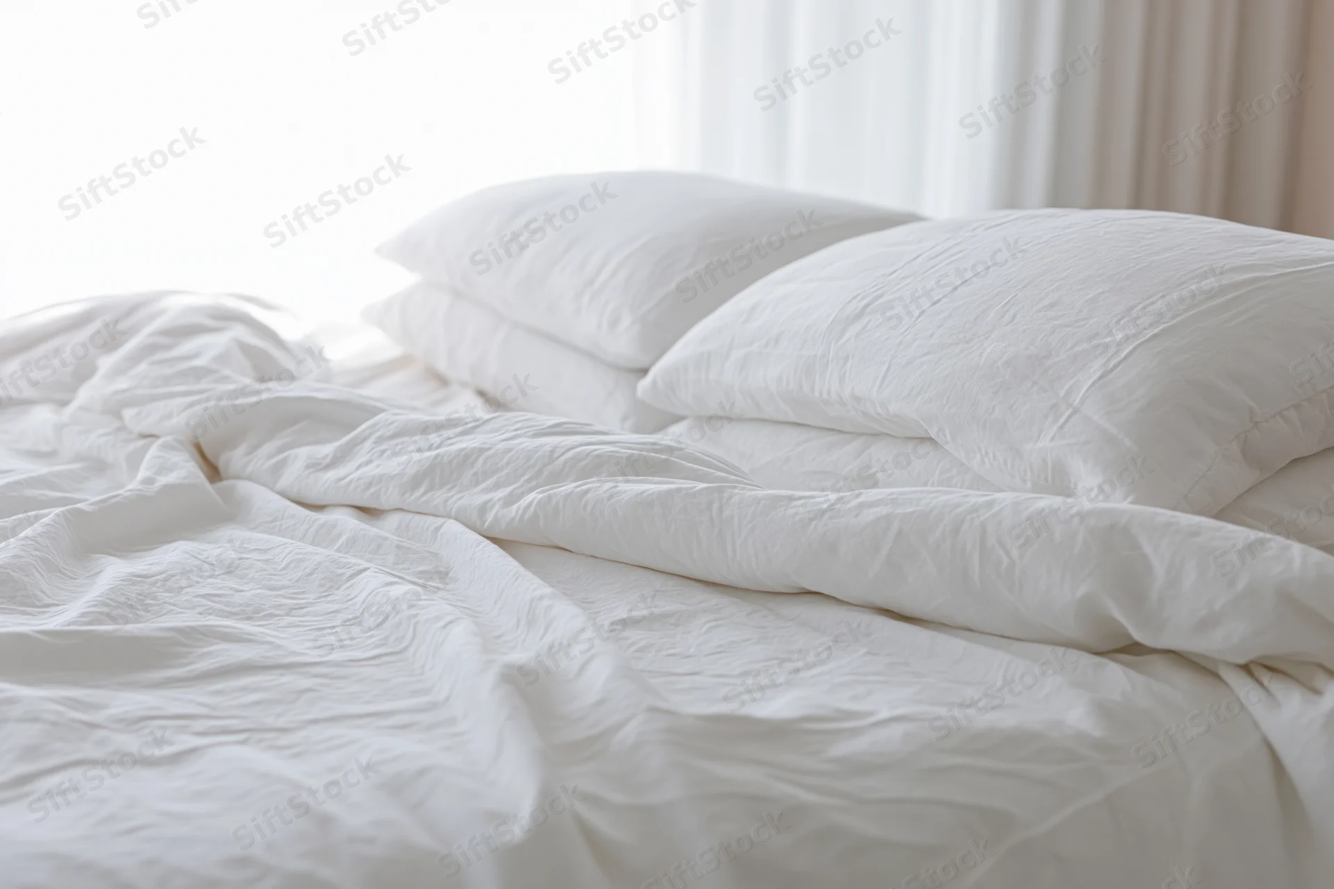 Unmade bed with rumpled white cotton duvet and stacked pillows in a hotel bedroom
