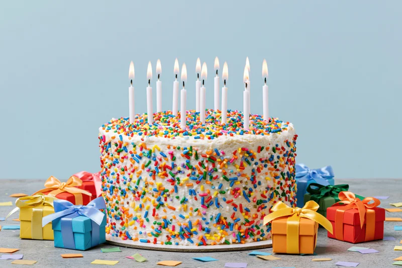 White-frosted birthday cake with rainbow sprinkles and lit candles surrounded by wrapped gifts and confetti on a table