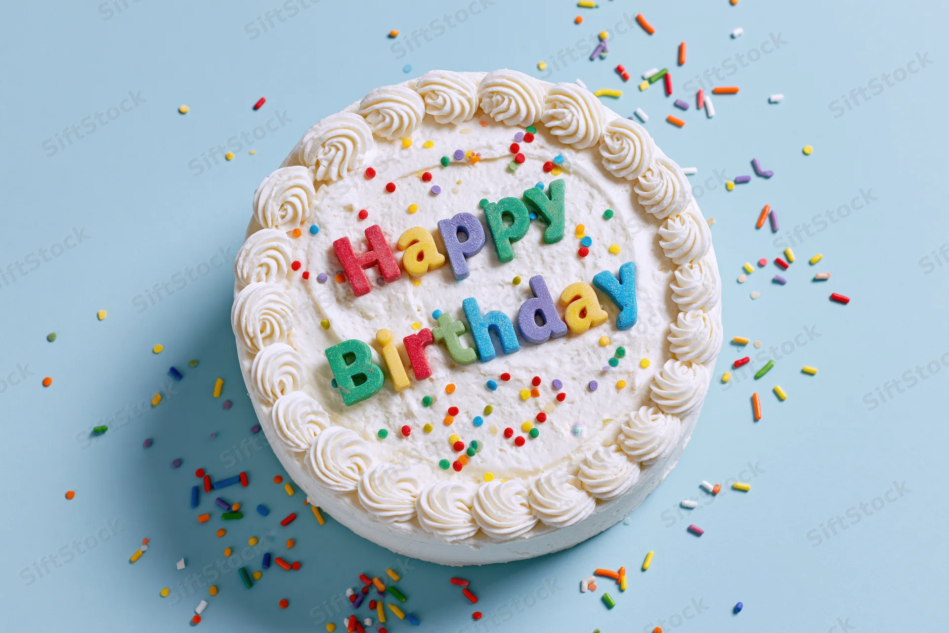 Round white-frosted birthday cake with colorful sprinkles and "Happy Birthday" letters on a blue surface