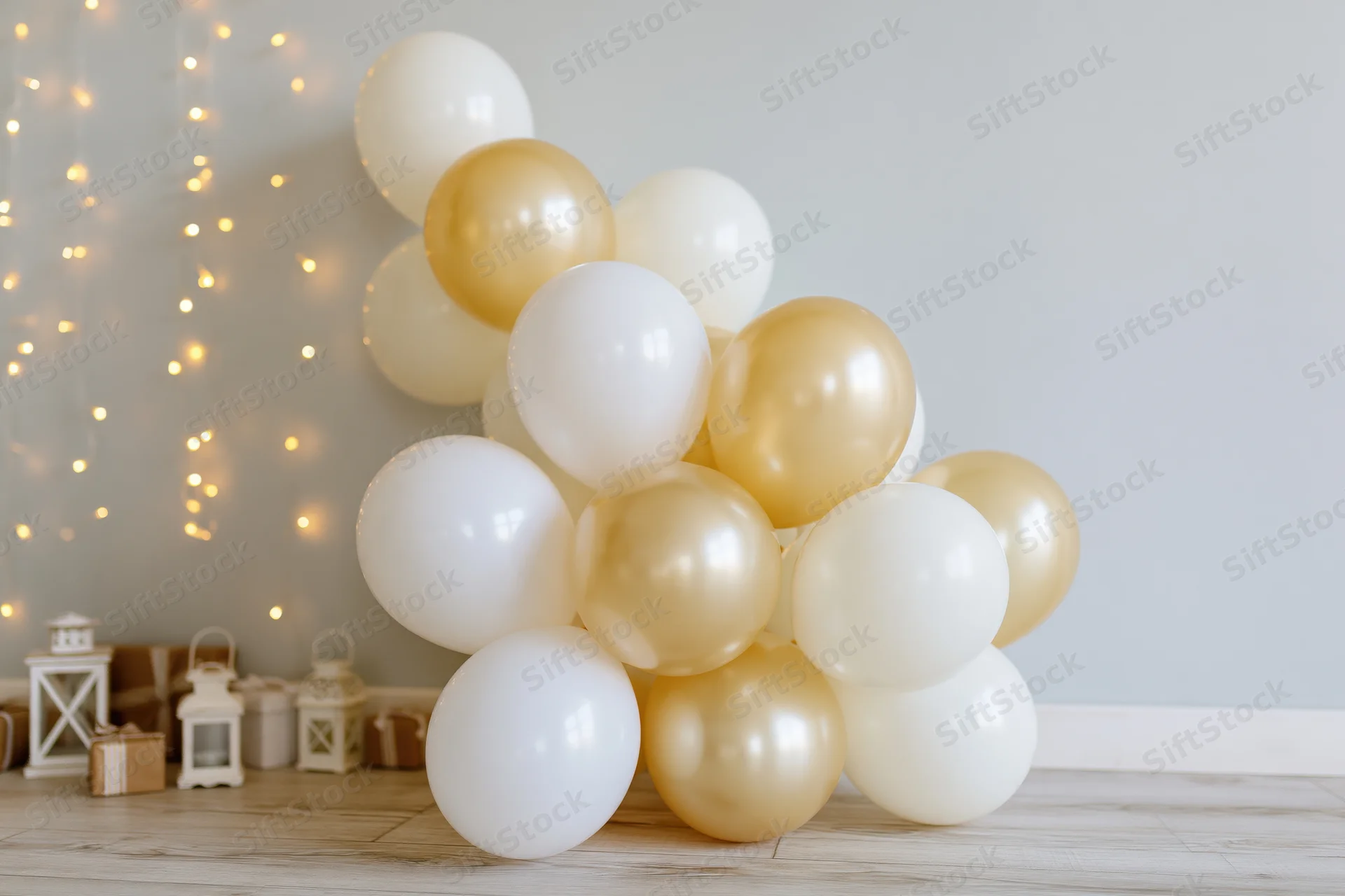 White and gold balloons clustered on wooden floor against pale grey wall with string lights and lanterns