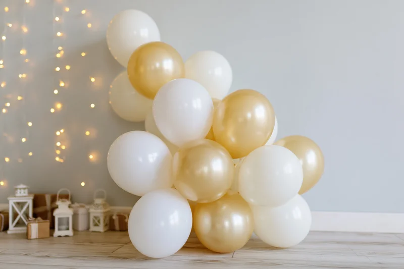 White and gold balloons clustered on wooden floor against pale grey wall with string lights and lanterns