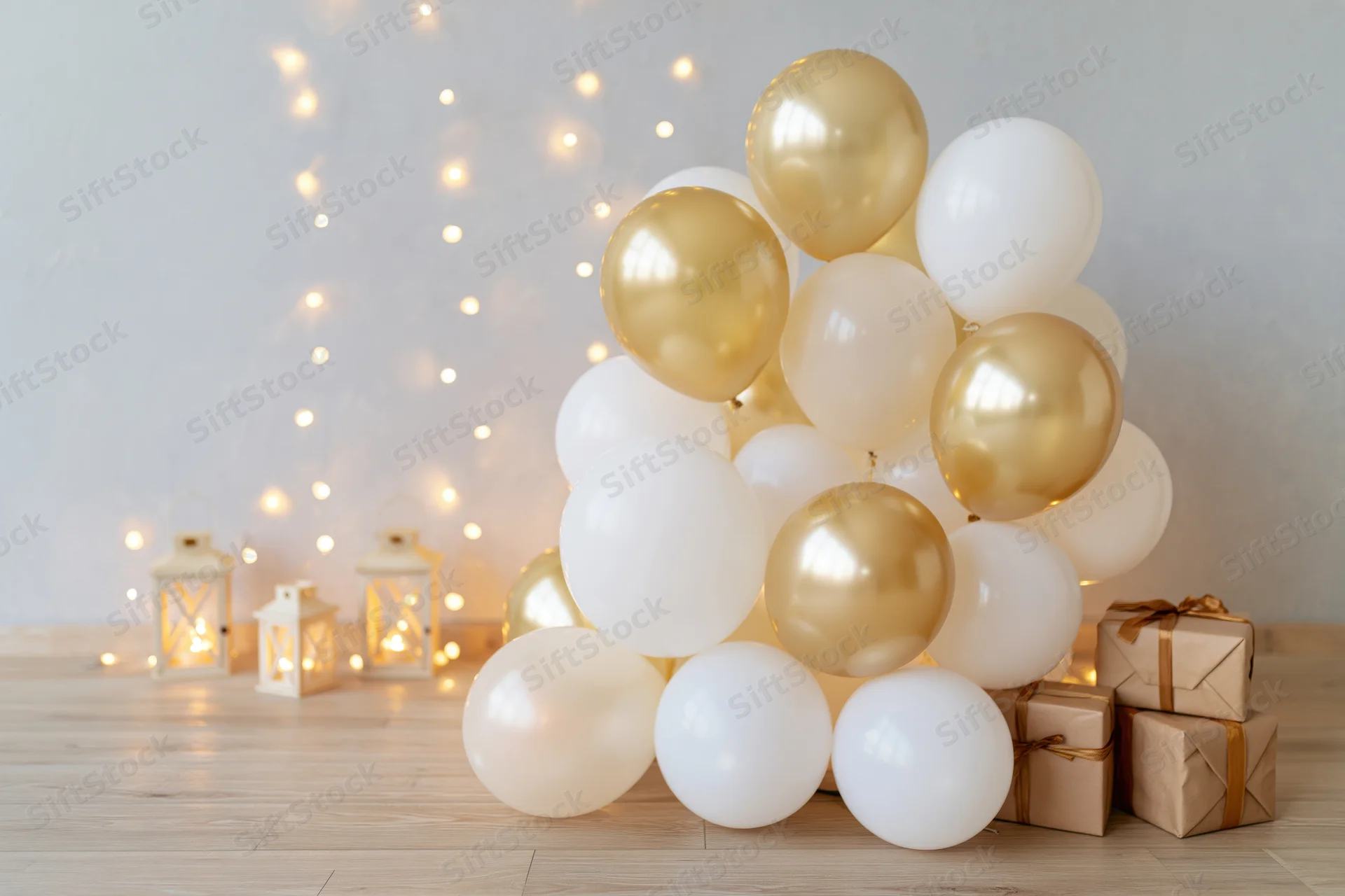 Cluster of white and gold balloons with wrapped gifts and glowing lanterns on wooden floor against pale grey wall