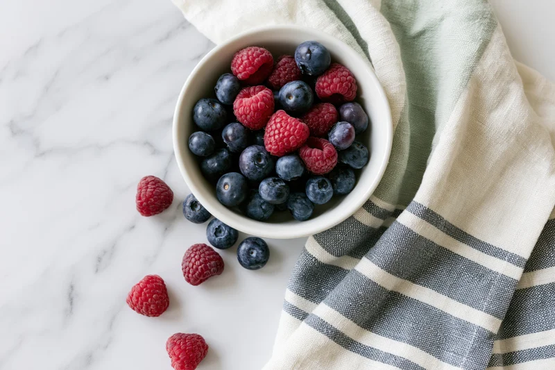 White ceramic bowl filled with blueberries and raspberries on marble countertop beside striped kitchen towel