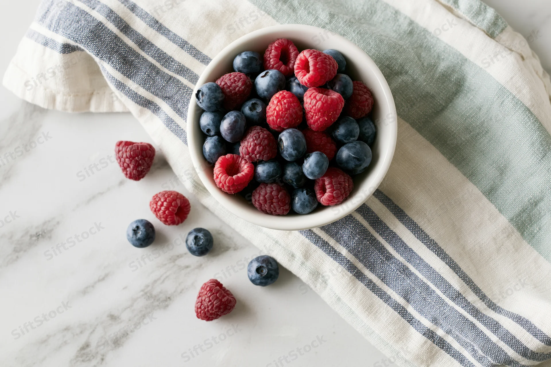 White ceramic bowl filled with fresh blueberries and raspberries on a striped cloth over marble countertop