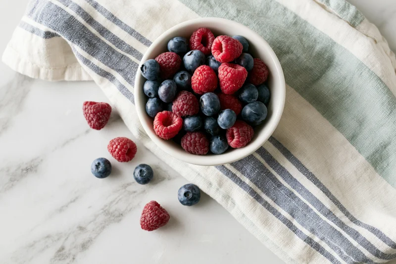 White ceramic bowl filled with fresh blueberries and raspberries on a striped cloth over marble countertop