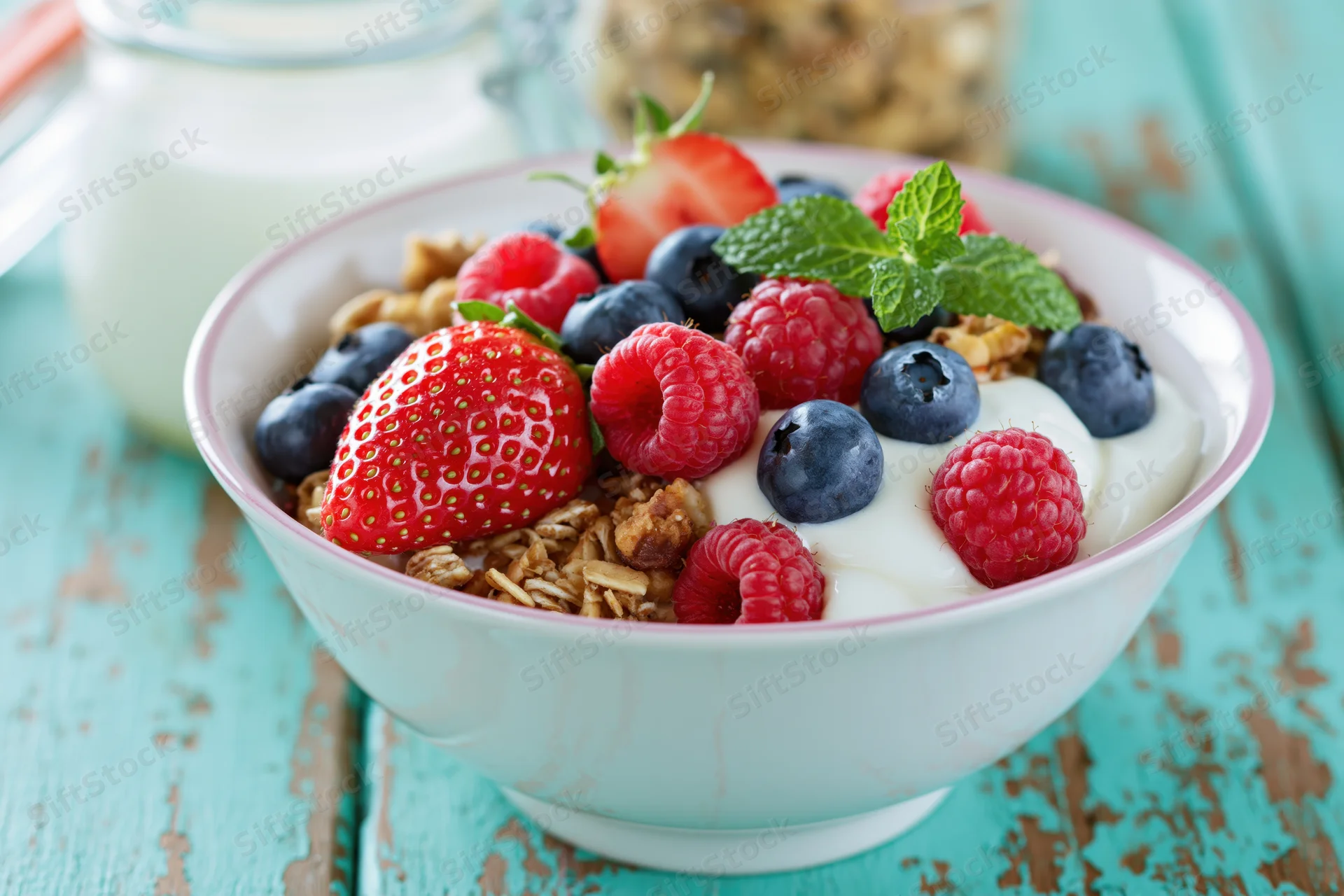 White ceramic bowl filled with granola, yogurt, strawberries, blueberries, raspberries, and mint on a turquoise wooden table