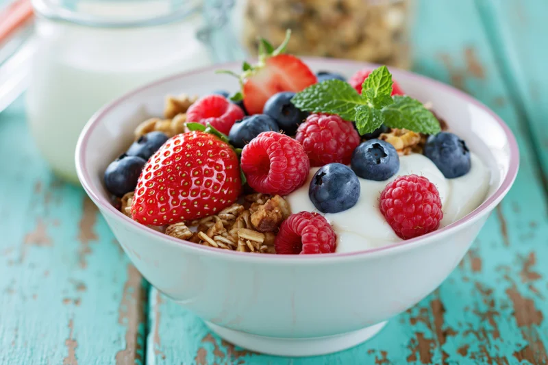 White ceramic bowl filled with granola, yogurt, strawberries, blueberries, raspberries, and mint on a turquoise wooden table