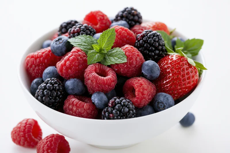 White ceramic bowl filled with mixed fresh berries and mint leaves on white background