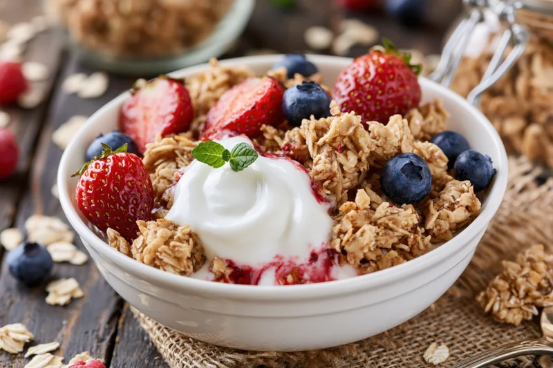 White ceramic bowl filled with granola, yogurt, strawberries and blueberries on wooden table