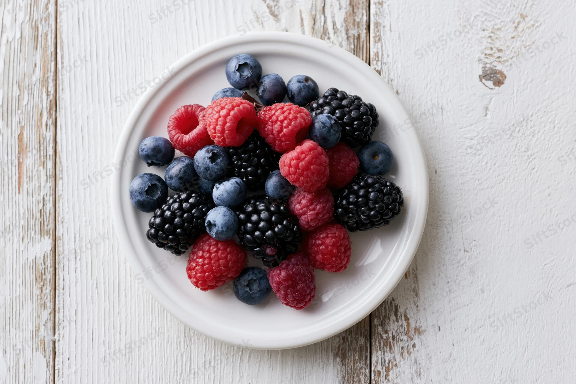 Fresh blueberries blackberries and raspberries on a white ceramic plate atop a weathered wooden surface