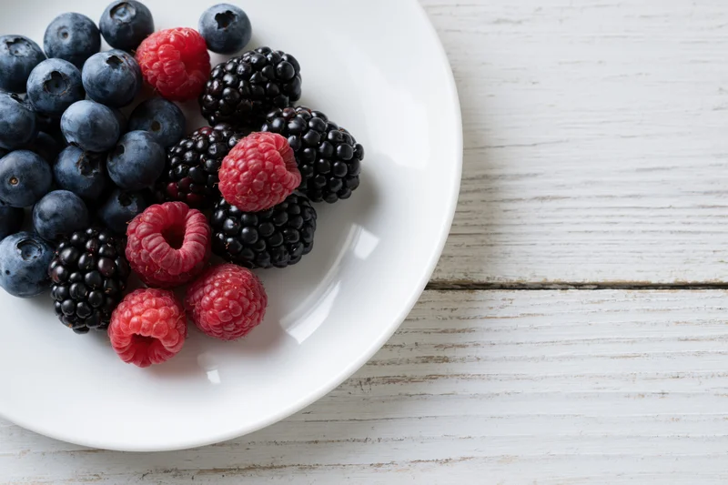 Fresh blueberries blackberries and raspberries on a white ceramic plate on a light wooden surface