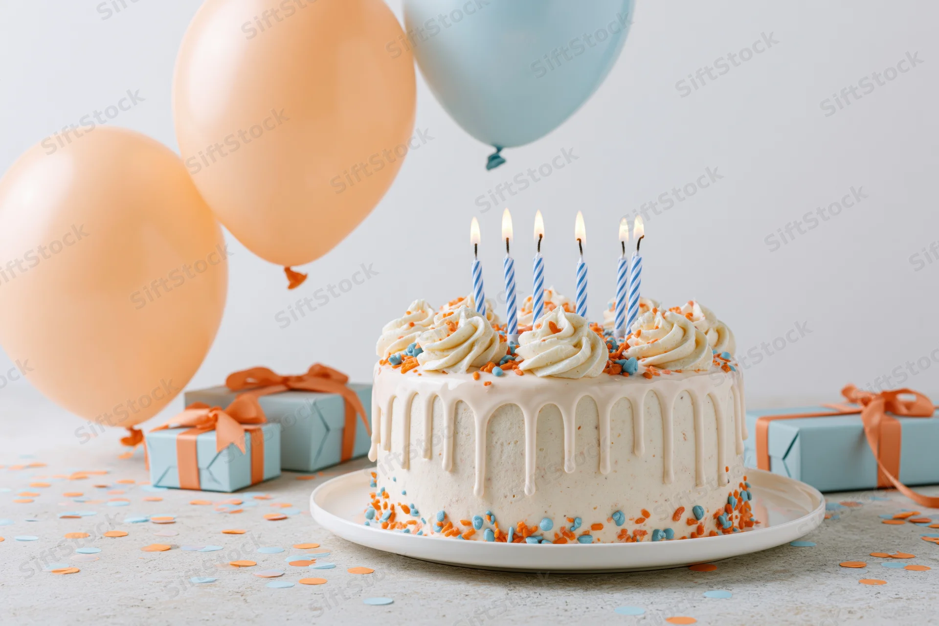 White drip birthday cake with lit striped candles on a table surrounded by balloons and gift boxes
