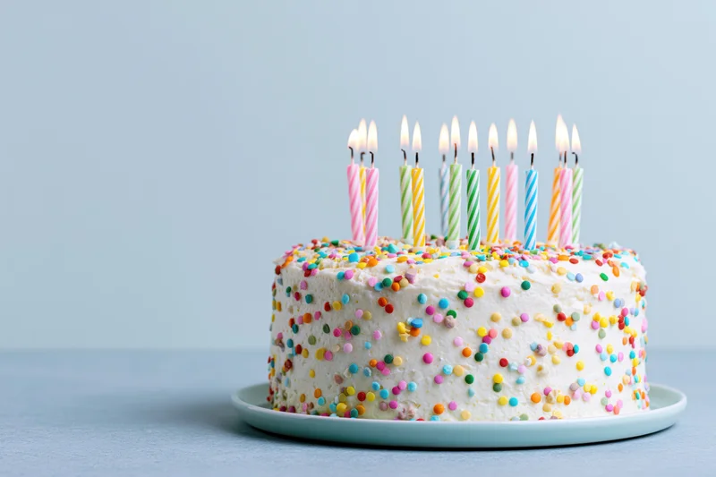 White frosted birthday cake with colorful sprinkles and lit candles on a plate against a light blue background