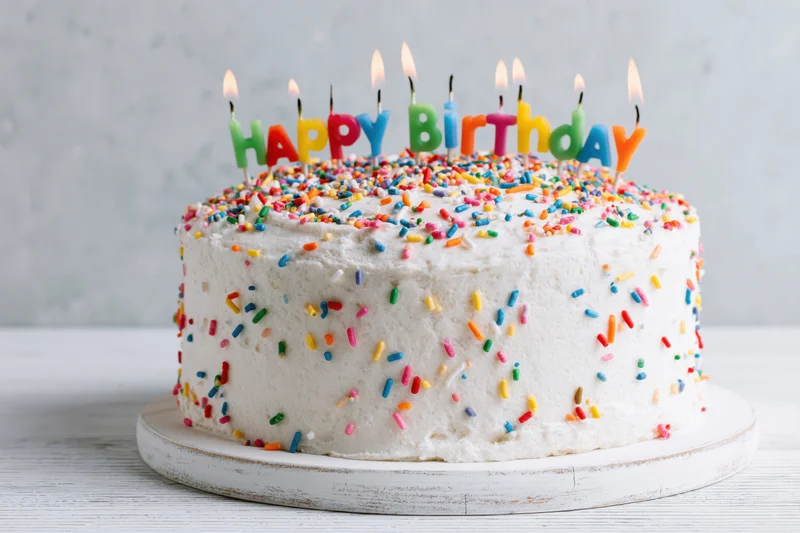 White frosted birthday cake with colorful sprinkles and lit candles spelling happy birthday on a wooden surface