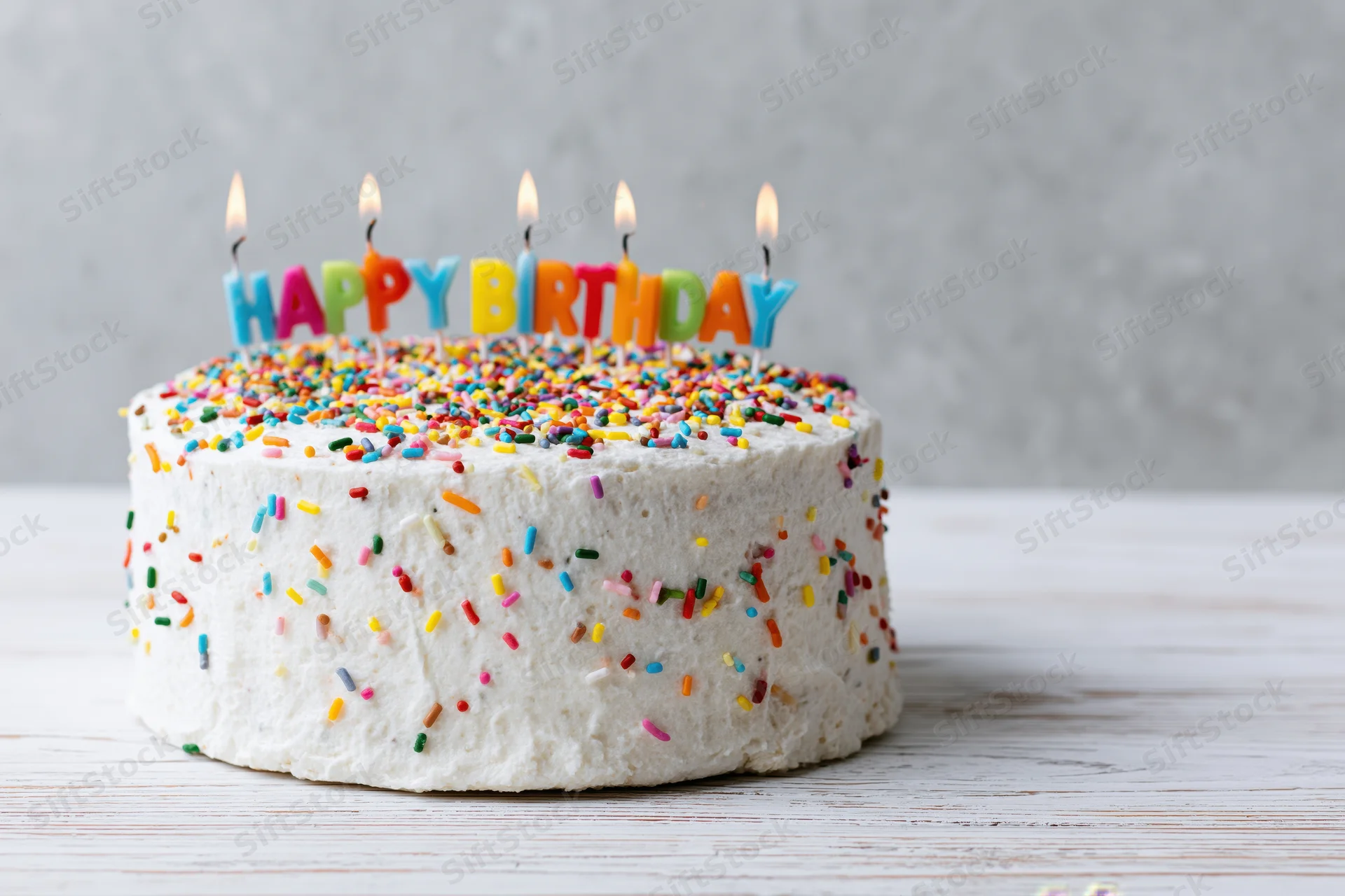 White frosted birthday cake with colorful sprinkles and lit candles spelling happy birthday on wooden table