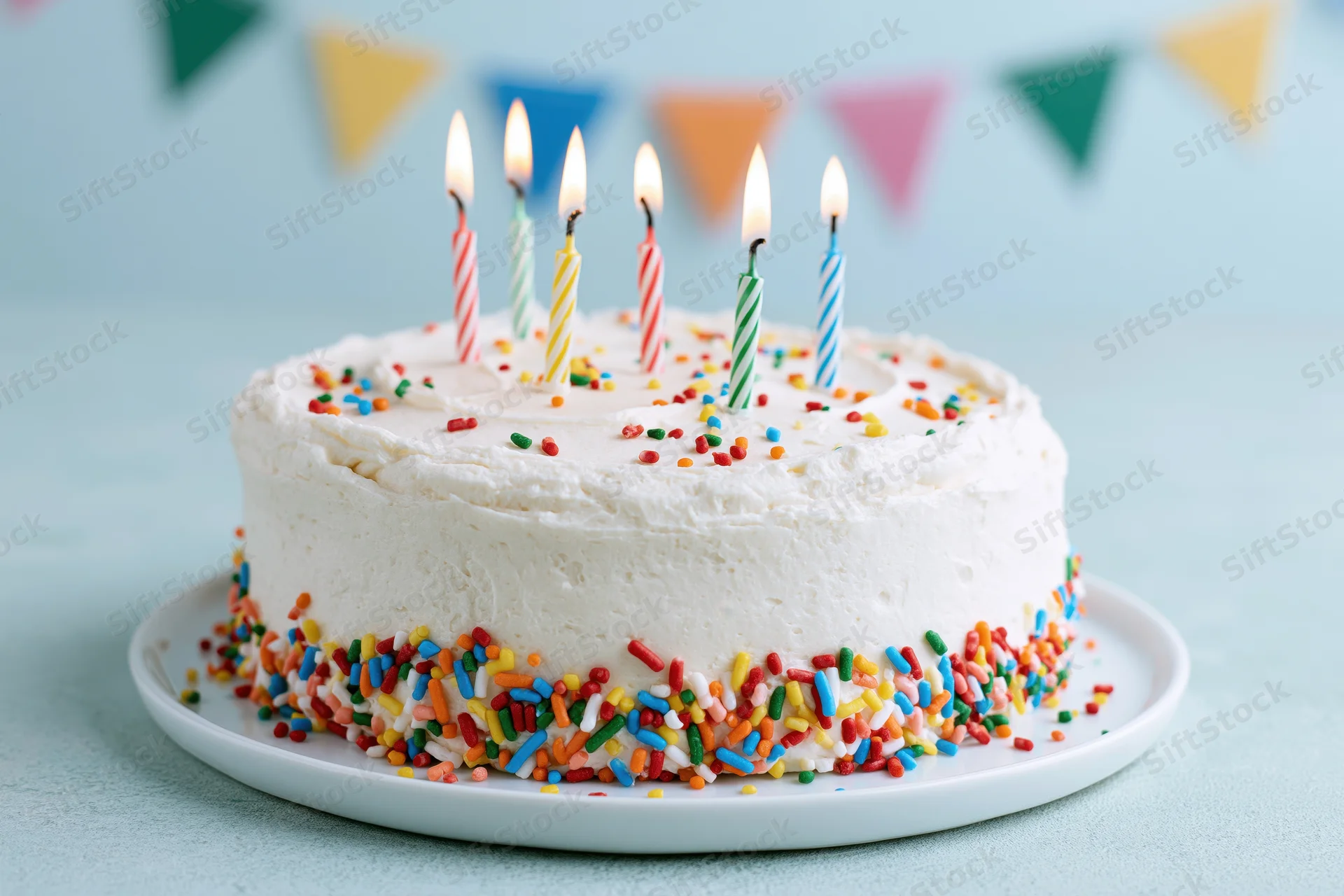 White frosted birthday cake with rainbow sprinkles and lit candles on a plate against a light blue background with party banner