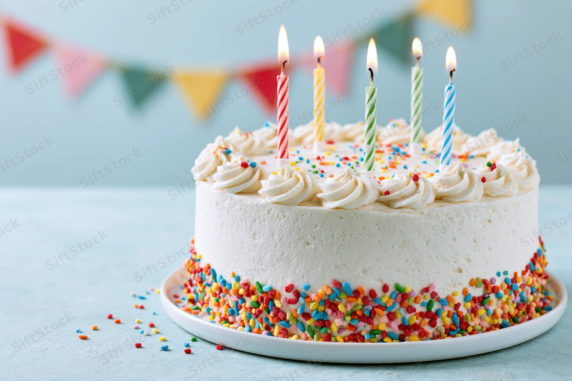 White frosted birthday cake with rainbow sprinkles and lit candles on a plate, set against a light blue background with colorful pennant banner