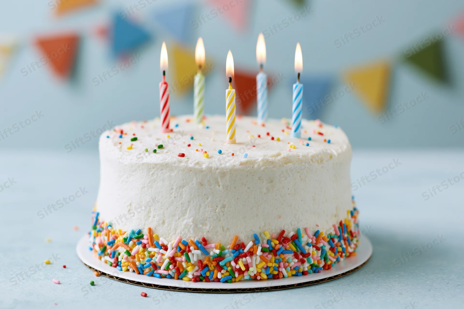 White frosted birthday cake with lit candles and rainbow sprinkles on a plate, set against a pastel blue background with colorful bunting in the distance