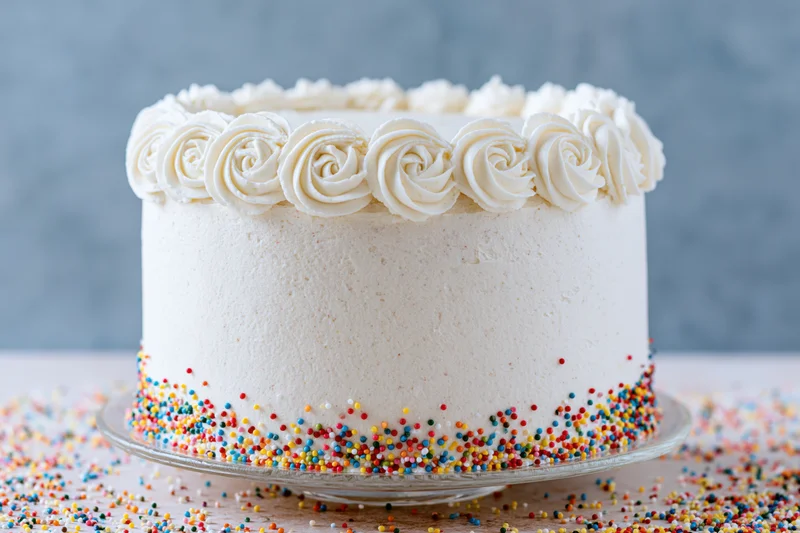 White frosted birthday cake with colorful sprinkles on a glass stand on wooden table