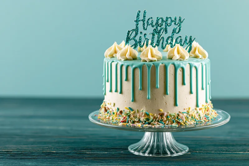 Birthday cake with teal drip frosting and sprinkles on a glass stand