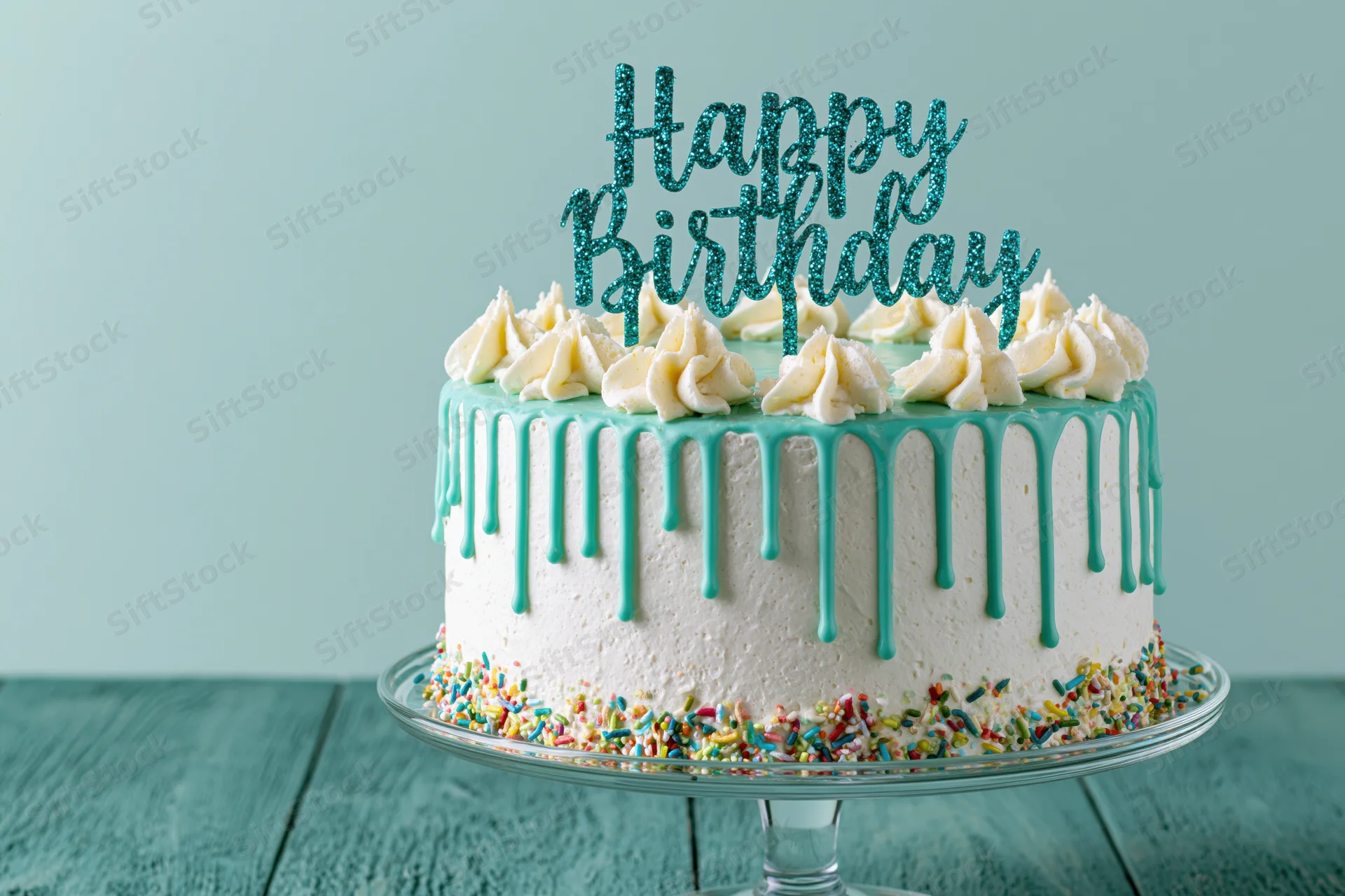White frosted birthday cake with teal drip icing and sprinkles on glass stand