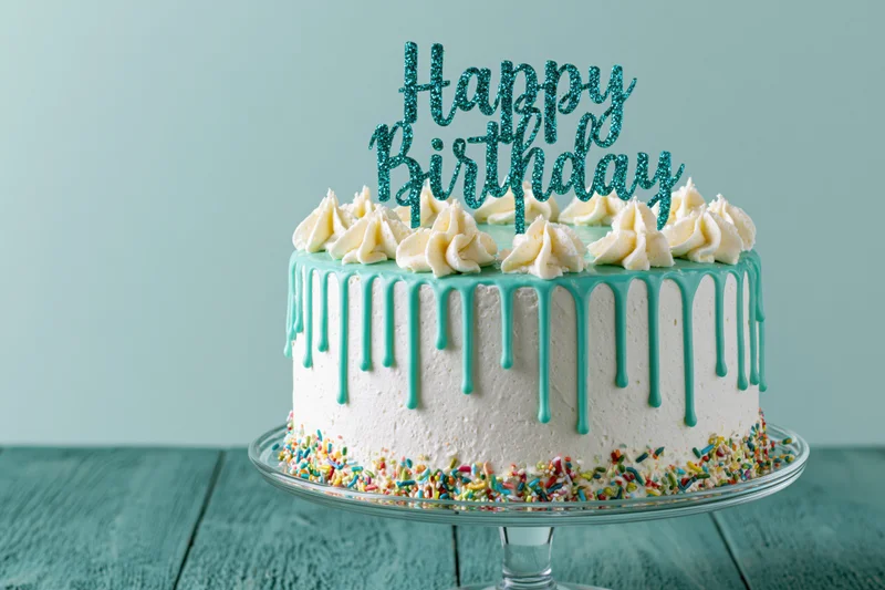 White frosted birthday cake with teal drip icing and sprinkles on glass stand