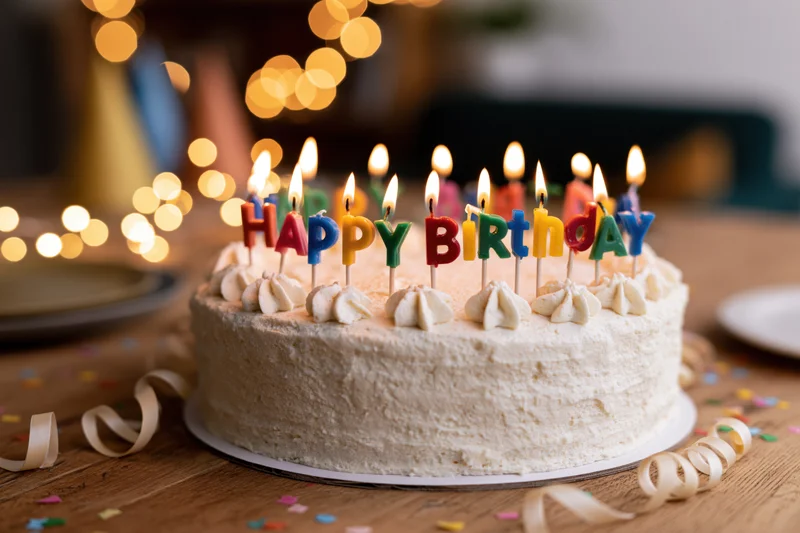 White frosted birthday cake with lit candles spelling happy birthday on wooden table surrounded by confetti and ribbon
