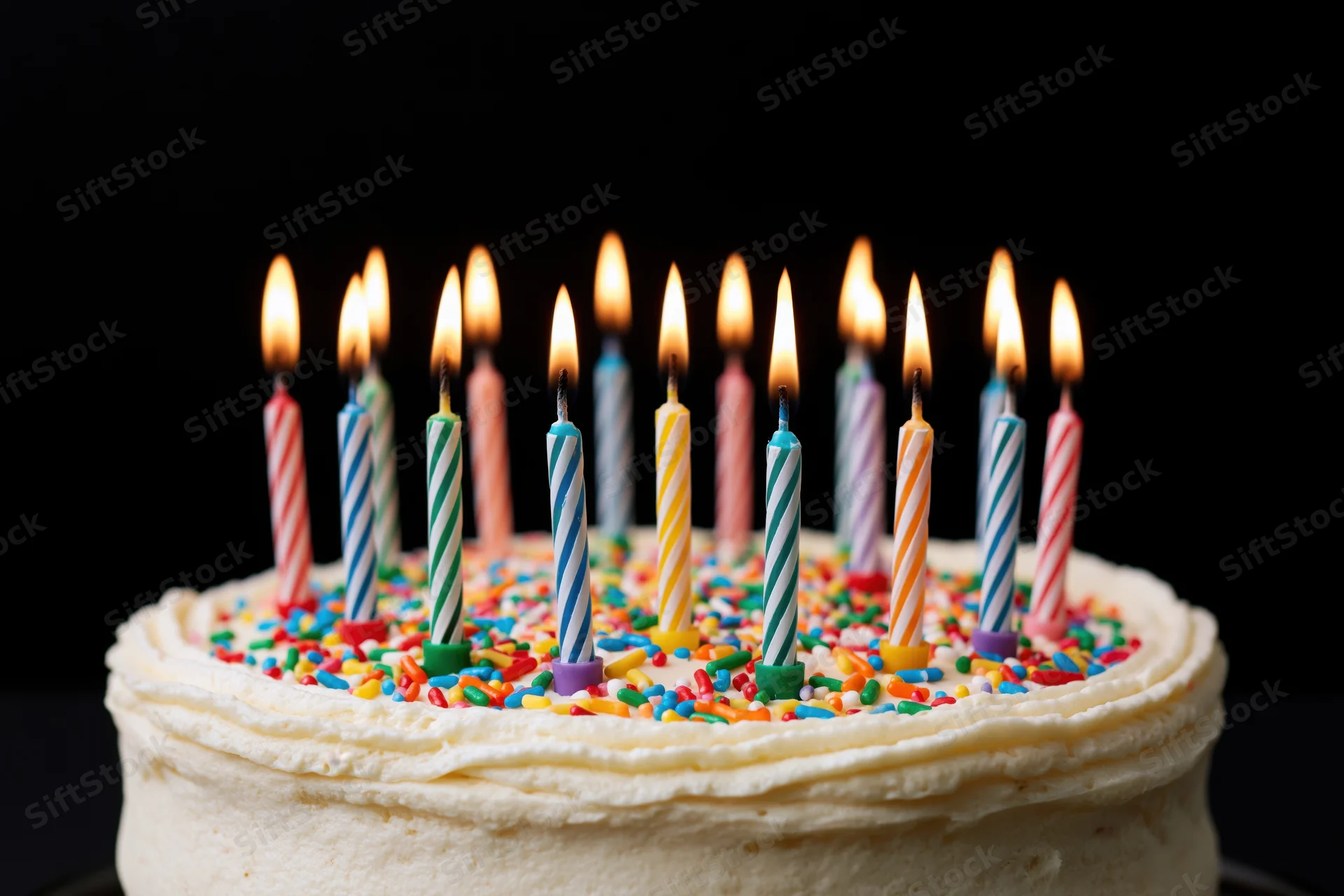 White frosted birthday cake with lit striped candles and colorful sprinkles on dark background