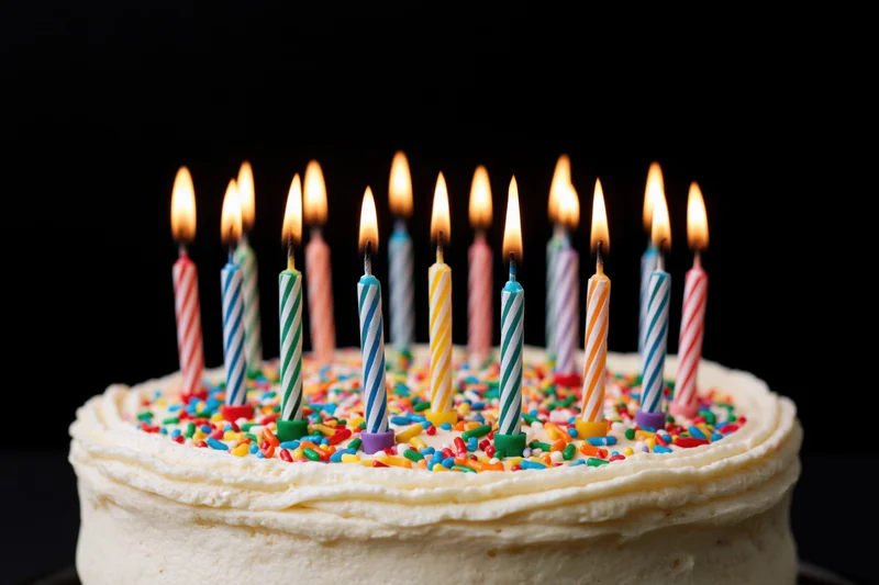 White frosted birthday cake with lit striped candles and colorful sprinkles on dark background