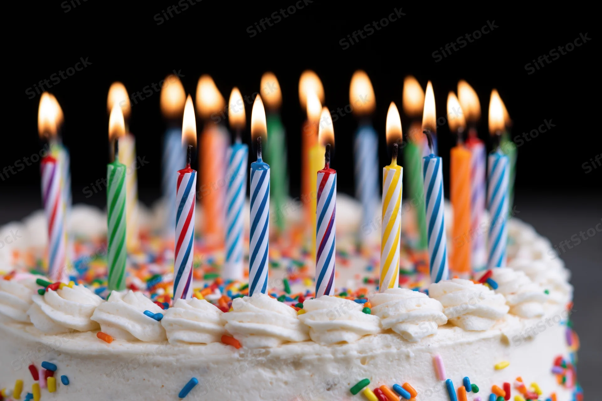 White frosted birthday cake with lit striped candles and sprinkles, close-up view