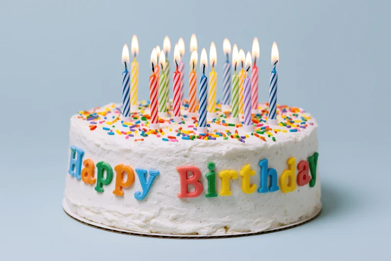 White frosted birthday cake with lit candles and colorful sprinkles, decorated with "Happy Birthday" lettering on side