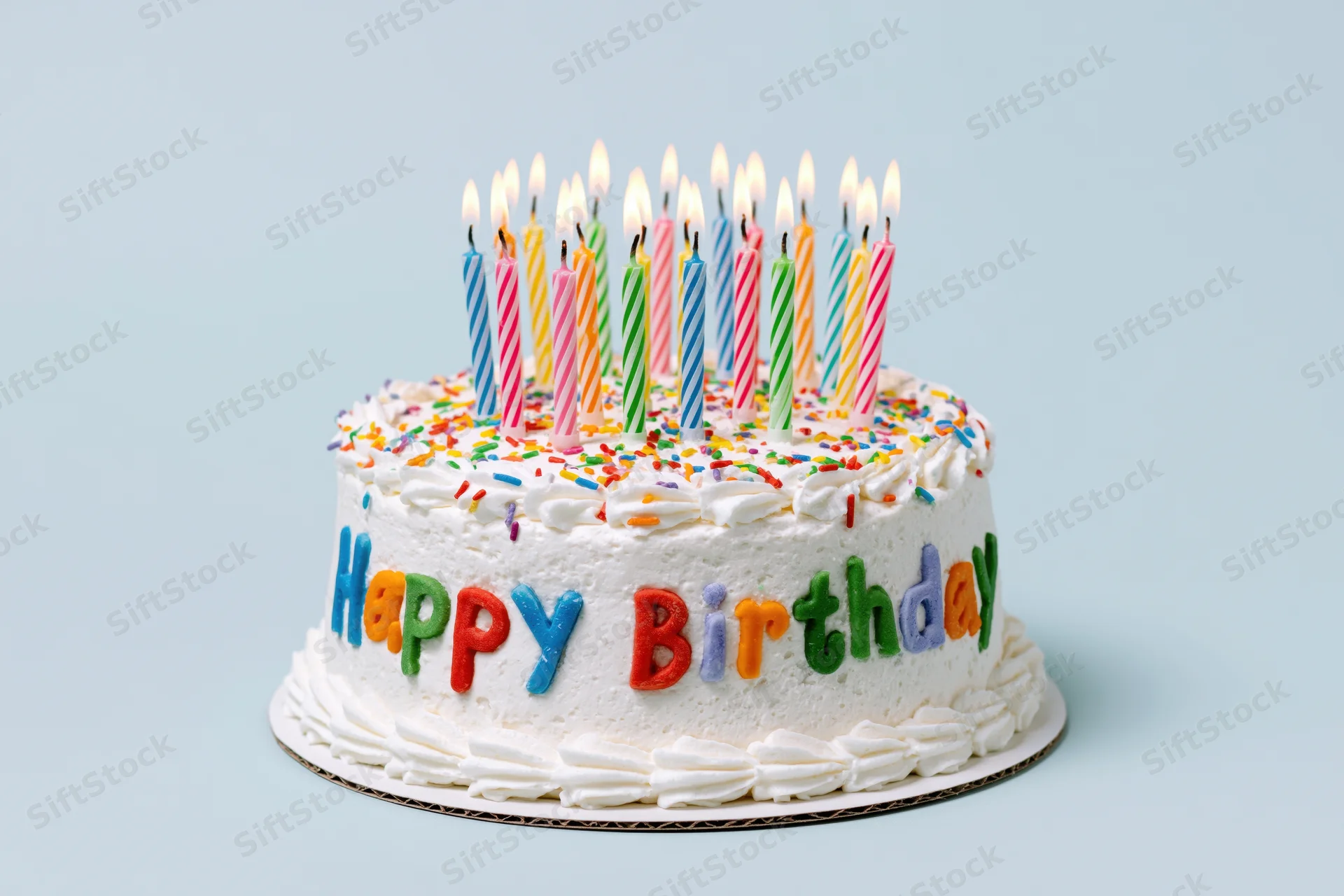 White frosted birthday cake with lit candles and colorful sprinkles, decorated with "Happy Birthday" letters on a light blue background