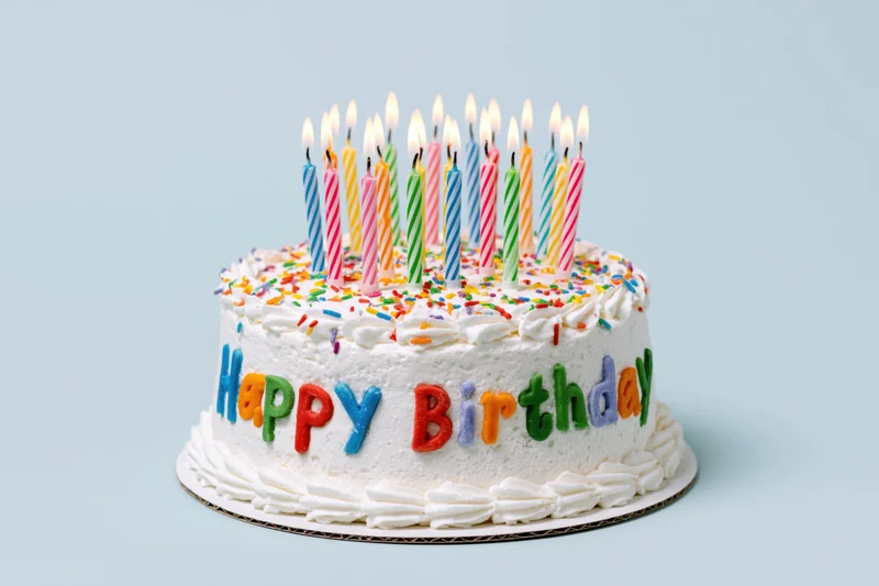 White frosted birthday cake with lit candles and colorful sprinkles, decorated with "Happy Birthday" letters on a light blue background