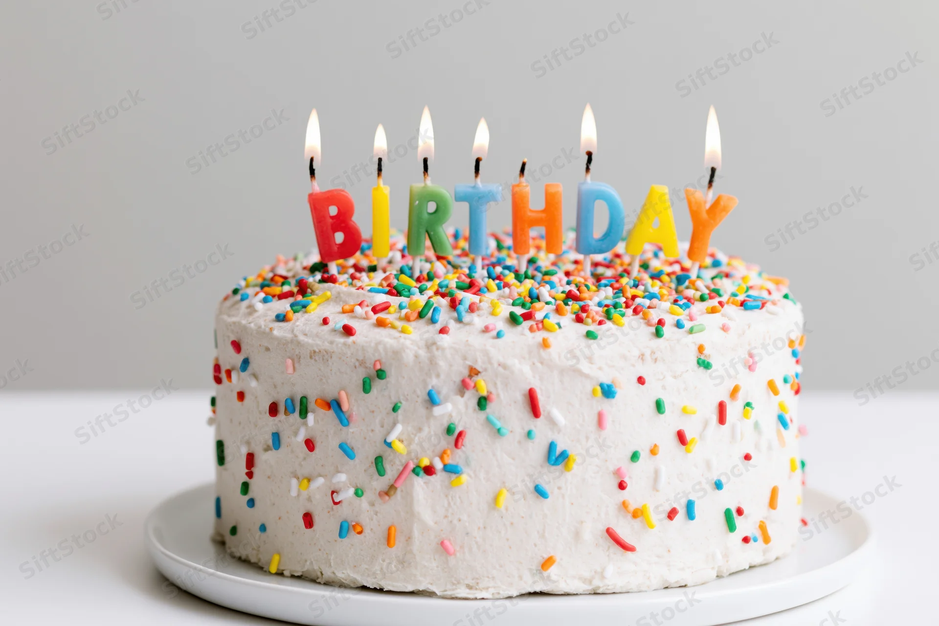 White frosted birthday cake with lit letter-shaped candles and colorful sprinkles on a plate