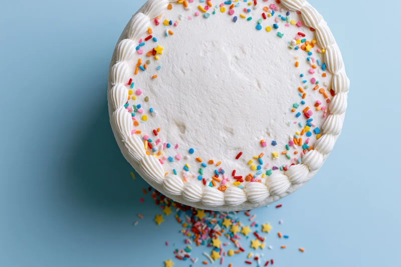 White frosted birthday cake with piped shell border and colorful sprinkles on blue background