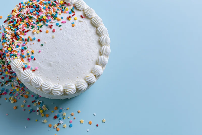 White frosted birthday cake with piped shell border and colorful sprinkles on blue background