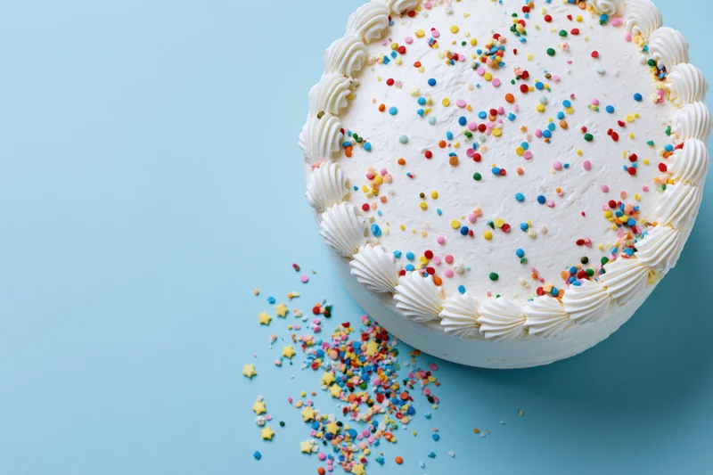 White frosted birthday cake with piped shell border and colorful sprinkles on blue surface