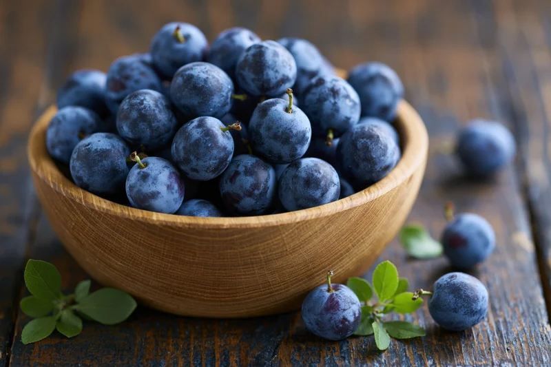 Wooden bowl filled with fresh sloe berries on a dark rustic table with scattered berries and green leaves