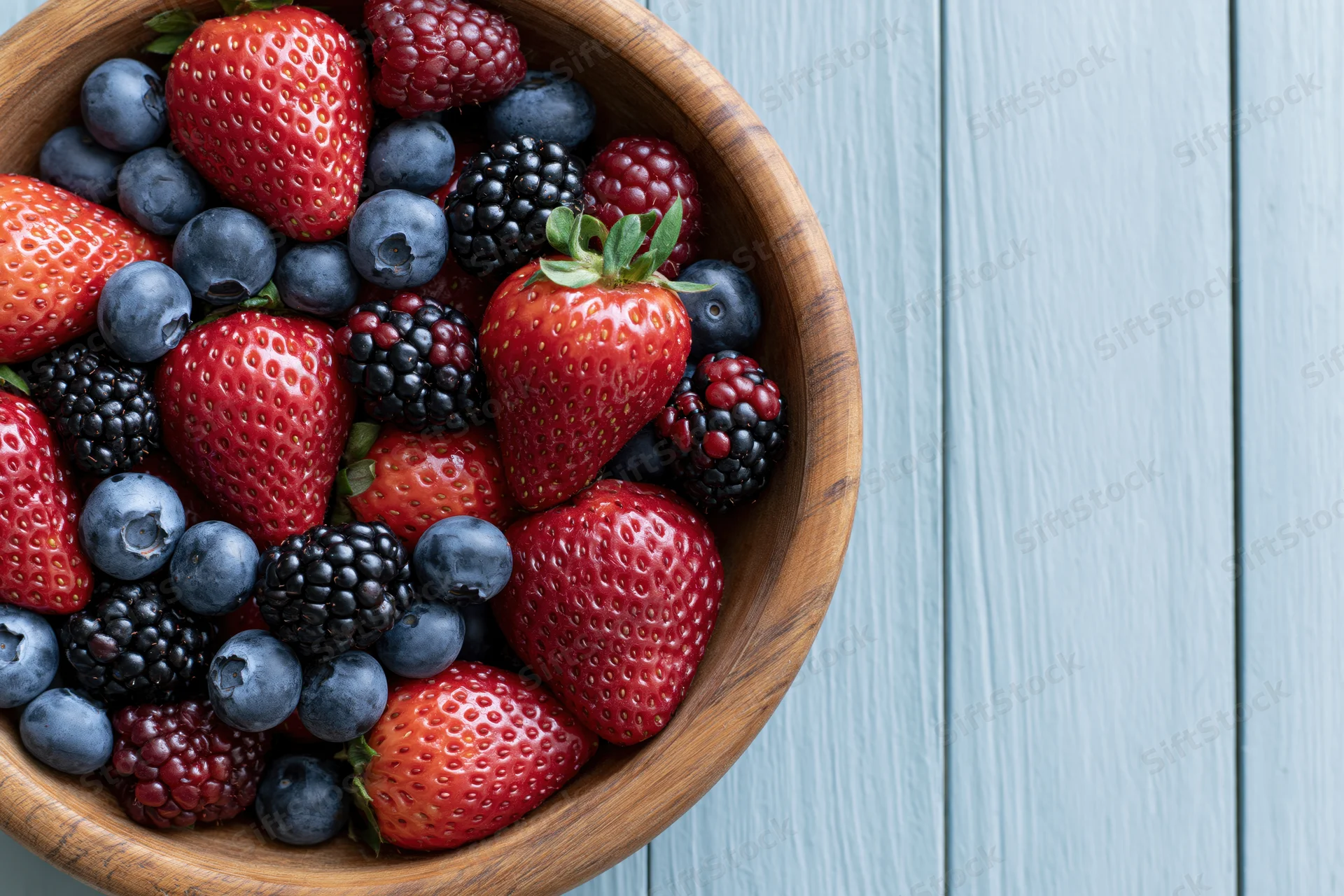 Wooden bowl filled with strawberries blueberries blackberries and raspberries on a blue wooden surface