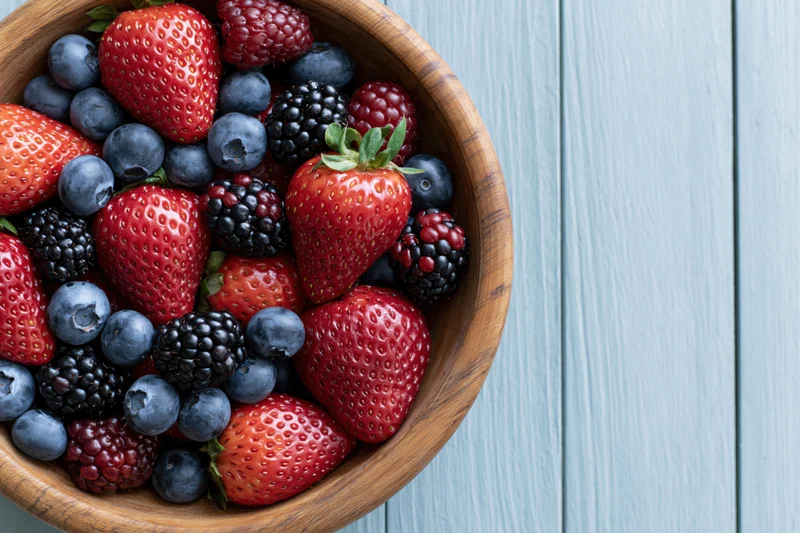 Wooden bowl filled with strawberries blueberries blackberries and raspberries on a blue wooden surface