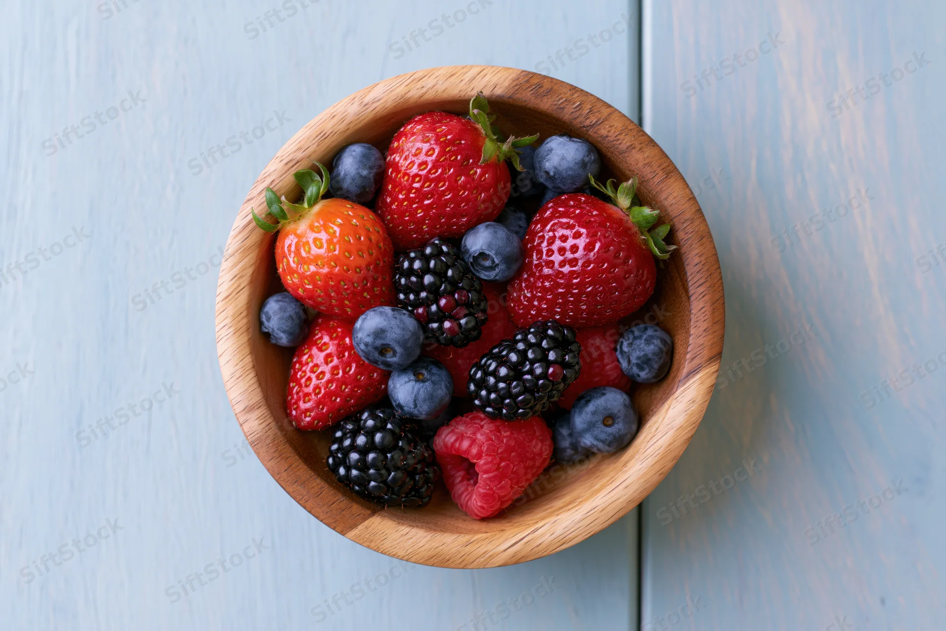 Wooden bowl filled with strawberries blueberries blackberries and raspberries on a blue wooden surface