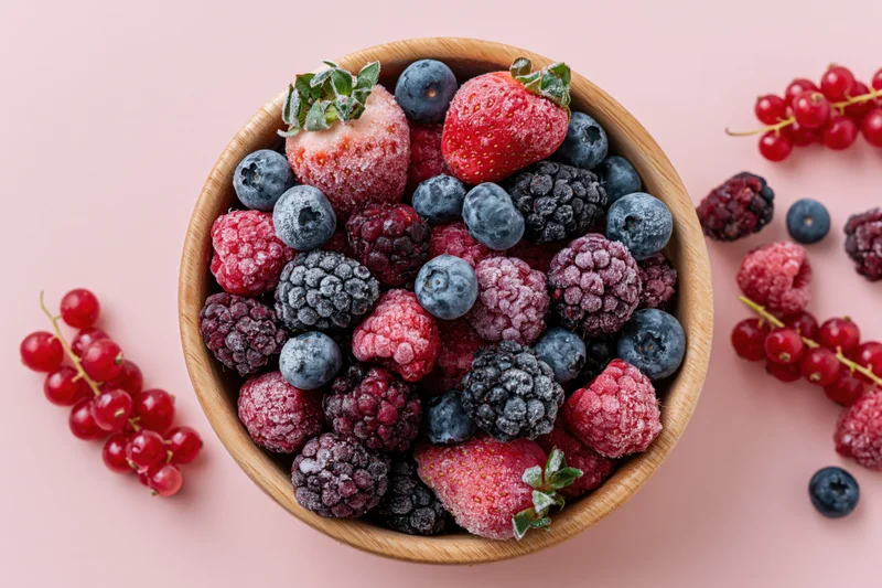 Frost-covered mixed berries in a wooden bowl on a pink surface