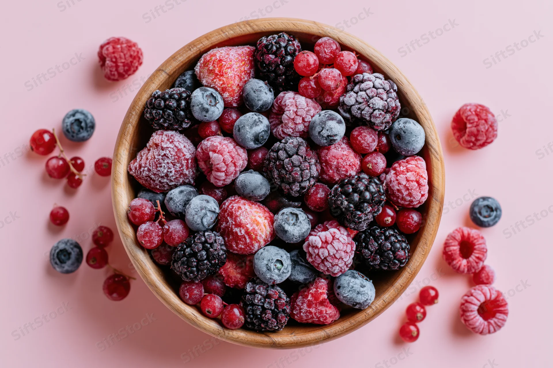 Wooden bowl filled with frost-covered frozen mixed berries on pink surface