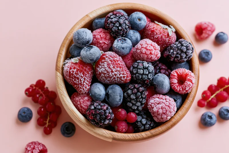 Wooden bowl filled with frost-covered frozen mixed berries on pink surface