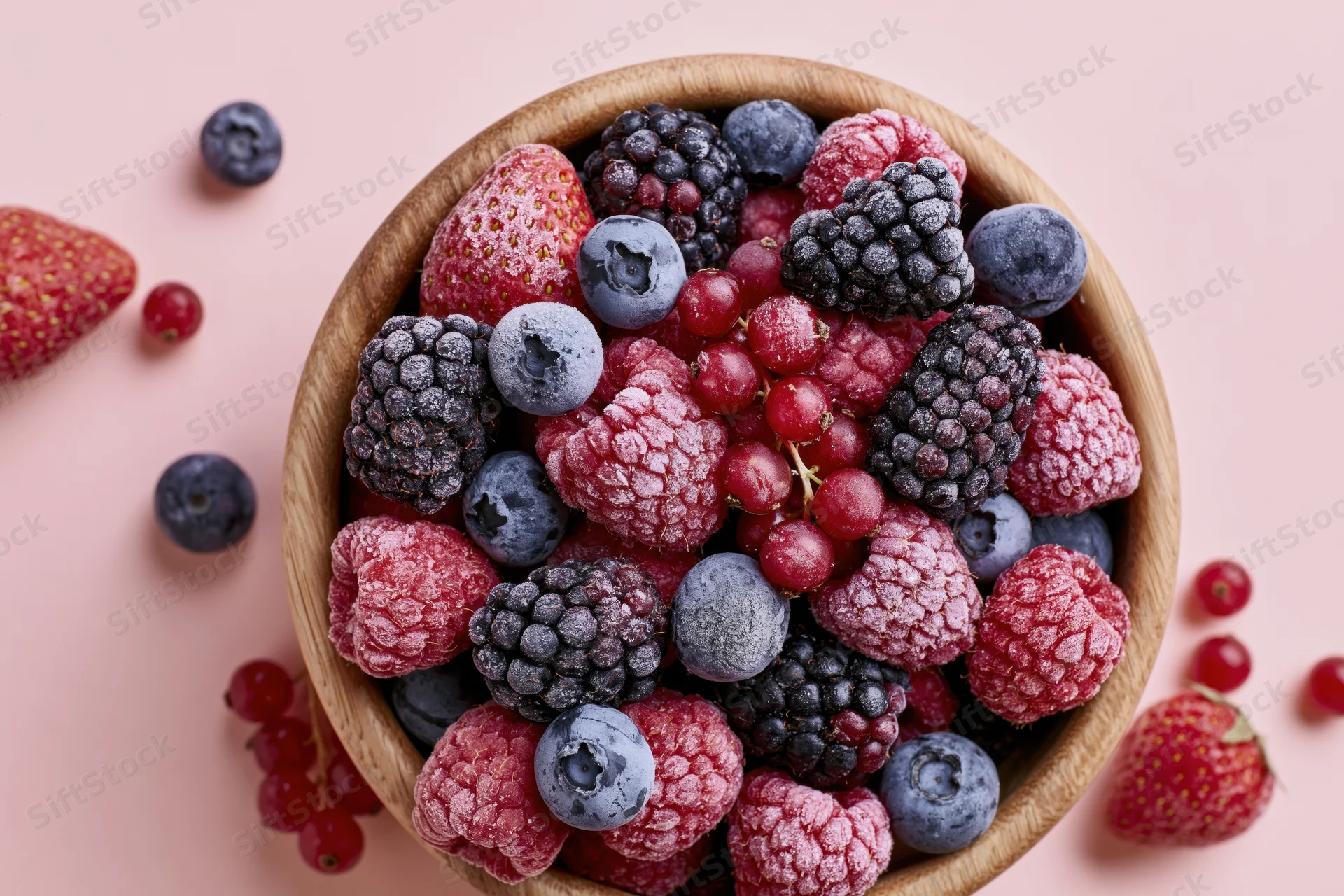 Wooden bowl filled with frost-covered frozen mixed berries on pink surface