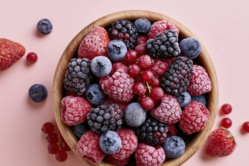 Wooden bowl filled with frost-covered frozen mixed berries on pink surface