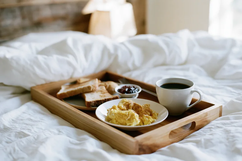Wooden tray with breakfast of scrambled eggs, toast, jam, and coffee on a bed