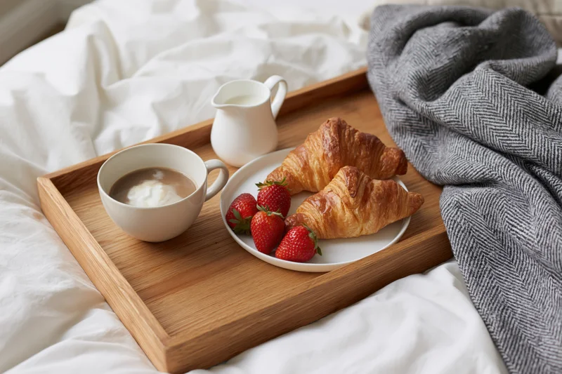 Wooden breakfast tray with croissants, coffee and strawberries on a rumpled white bed
