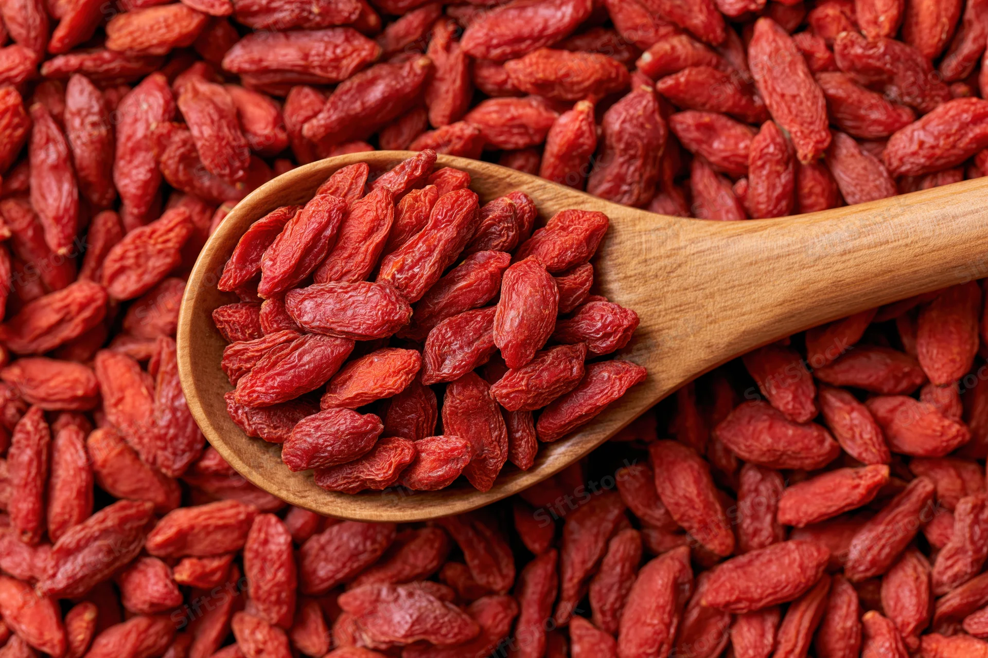 Wooden spoon filled with dried goji berries resting on a bed of the same berries in a flat lay arrangement