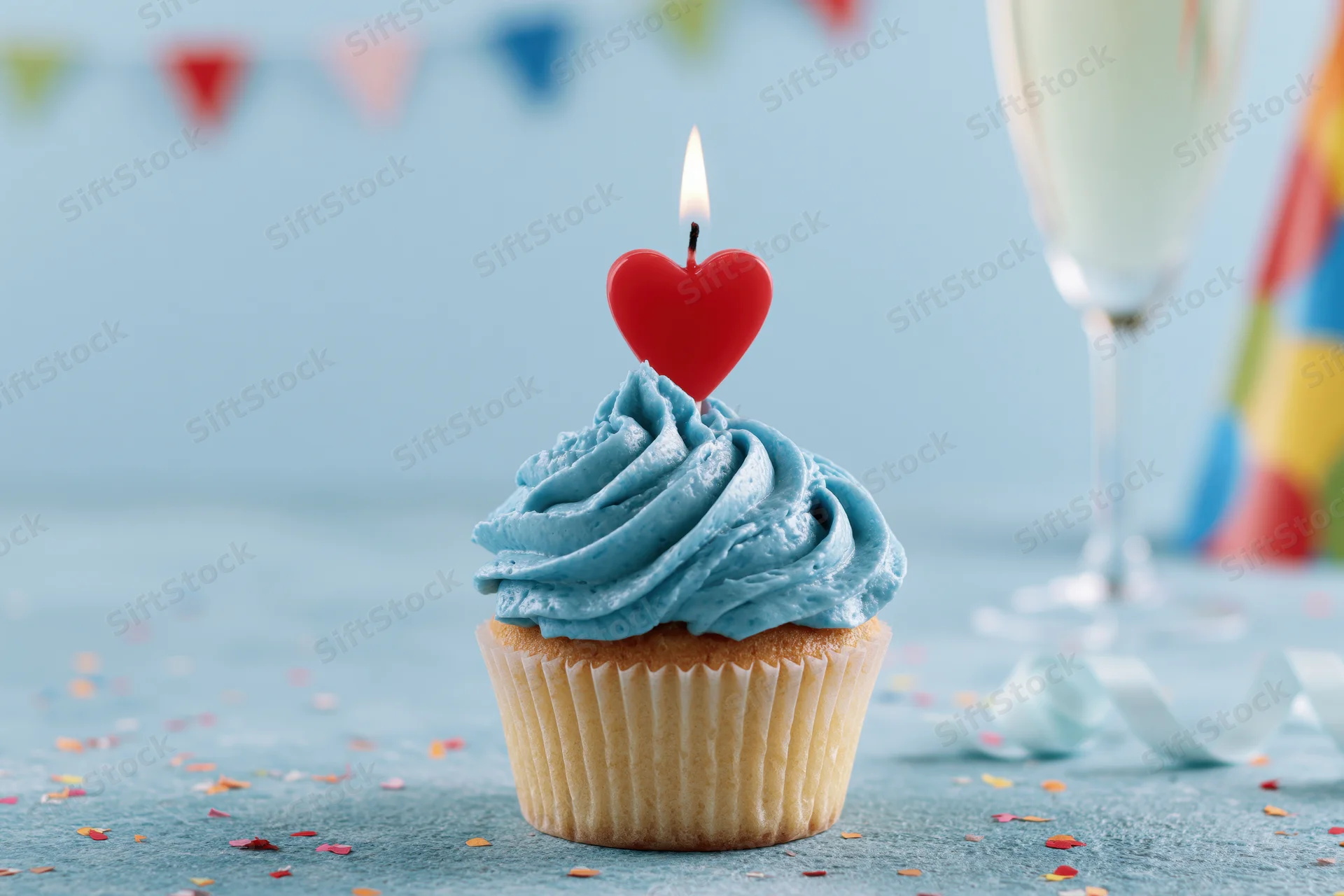 Birthday cupcake with blue frosting and lit red heart candle on a festive table with confetti and party decorations